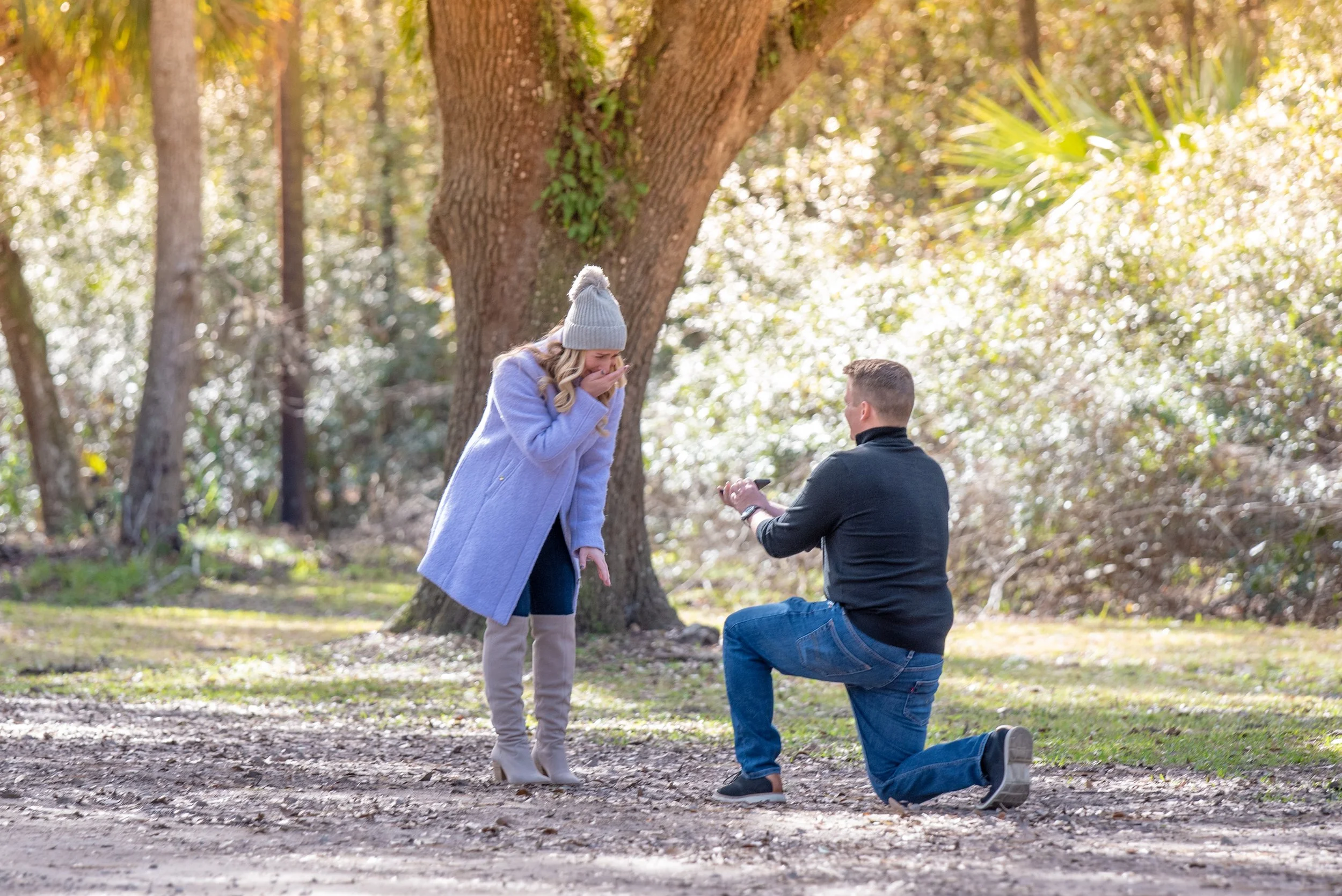 A man kneels on one knee in a park, presenting a ring to a surprised woman wearing a light blue coat and gray beanie.