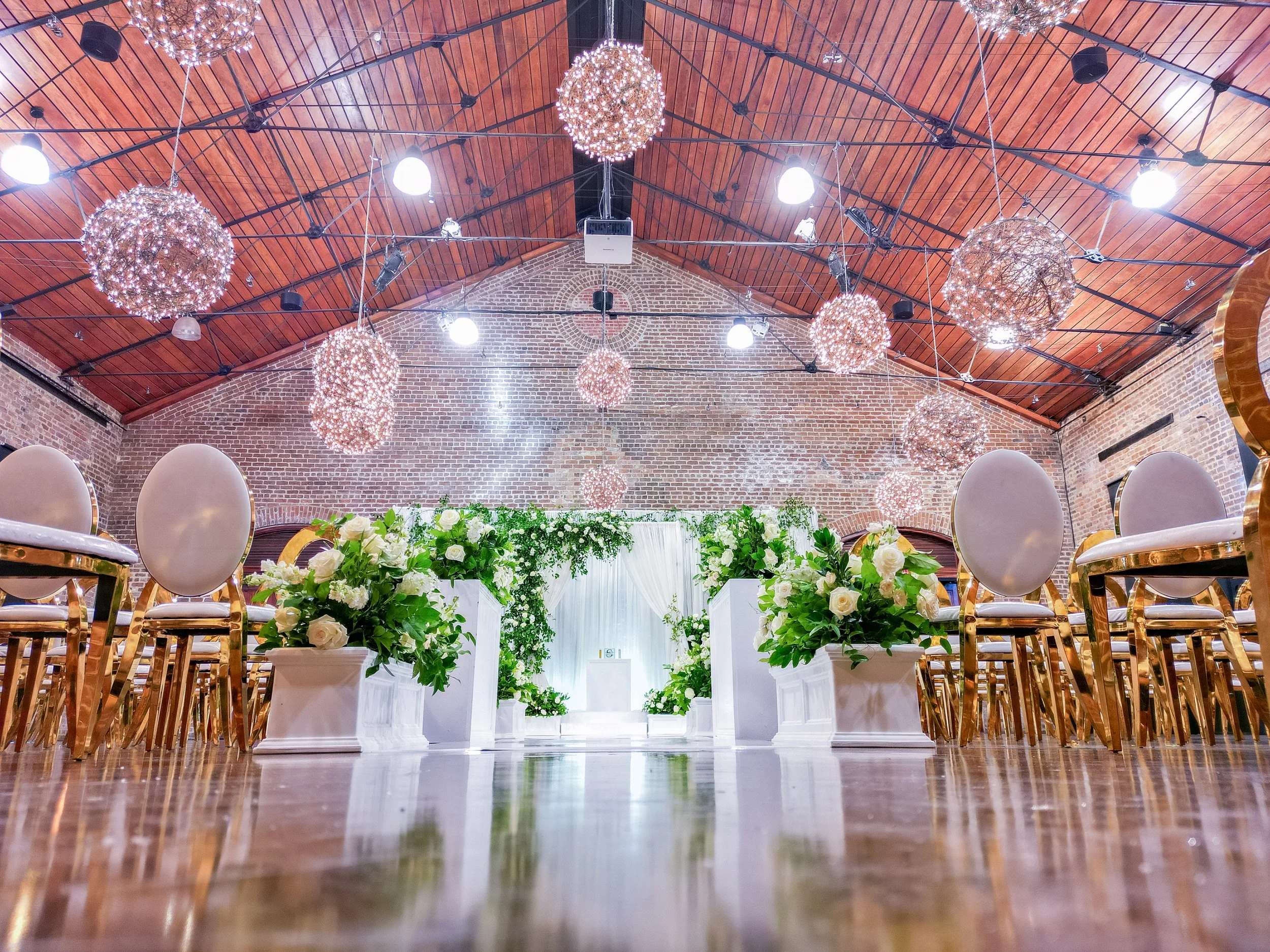 Wedding reception setup in a brick-walled hall with chandeliers, white roses, and gold chairs lining an aisle of greenery and flowers