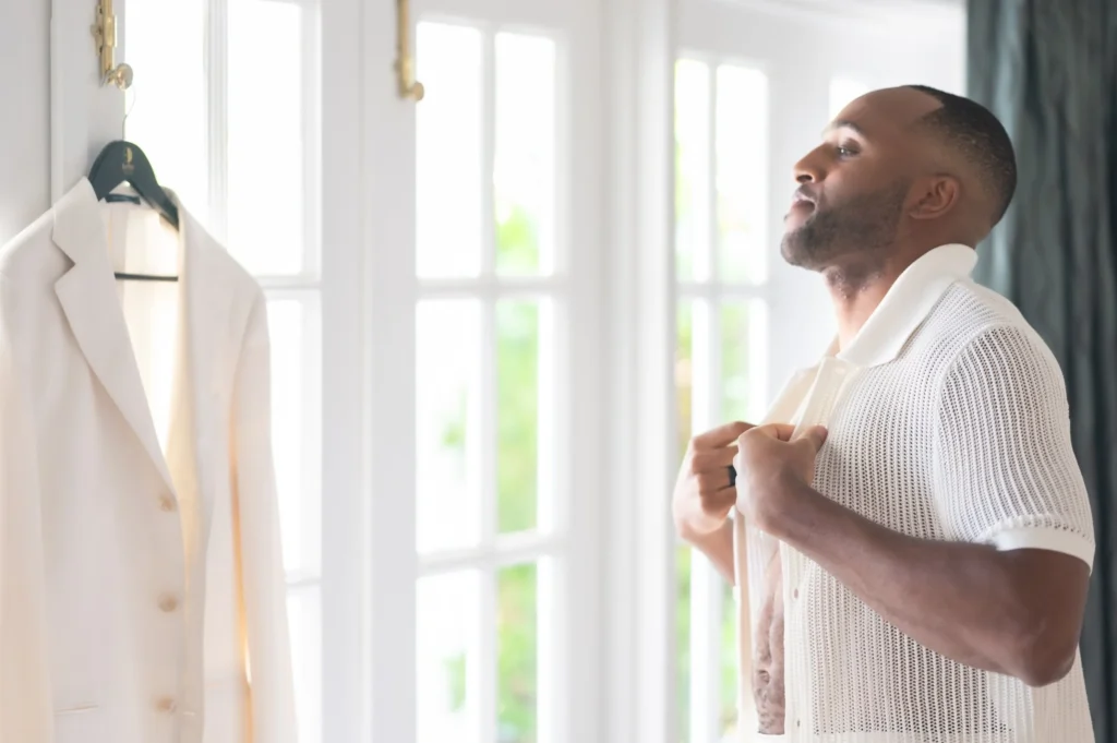 Arthur-Andrea-Jamaica-Halfmoon-Wedding-085.jpg - OMOS Photography Man wearing a white knit cardigan stands by a bright window, adjusting a white blazer on a hanger.
