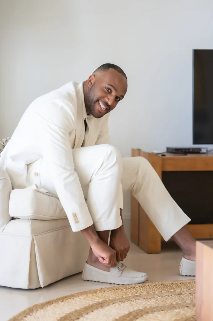 Arthur-Andrea-Jamaica-Halfmoon-Wedding-097.jpg - OMOS Photography Man in a cream suit bends down to tie a white loafer in a bright living room.