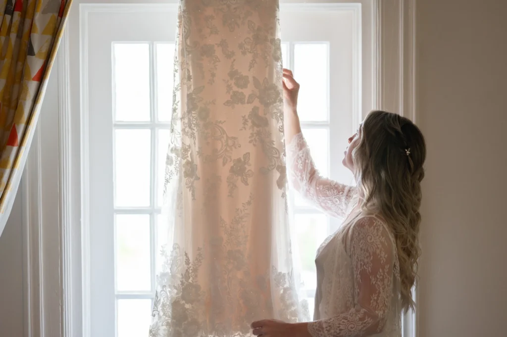 Arthur-Andrea-Jamaica-Halfmoon-Wedding-135.jpg - OMOS Photography Bride in a lace wedding dress pulls back a sheer lace curtain, revealing a bright window behind her on a softly lit day