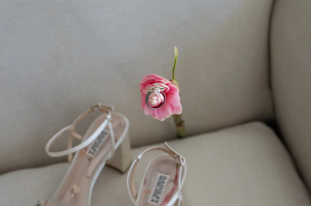 Arthur-Andrea-Jamaica-Halfmoon-Wedding-141.jpg - OMOS Photography Pink flower with an engagement ring and wedding band resting on its stem; blurred beige couch behind.