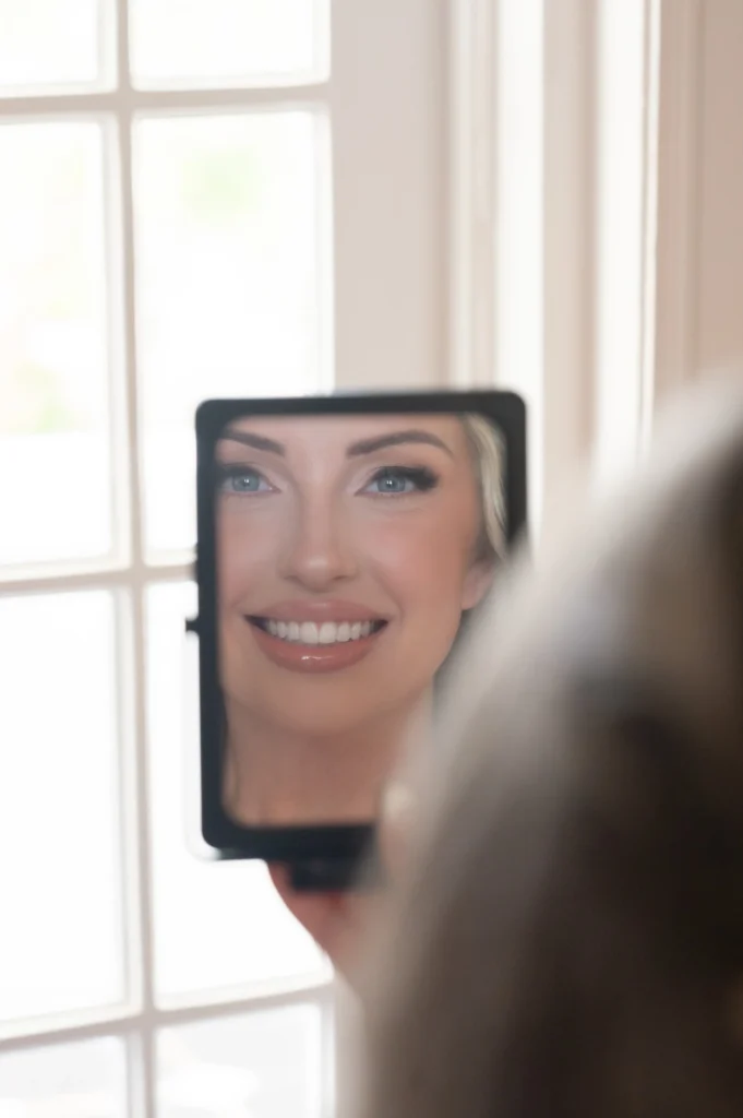 Arthur-Andrea-Jamaica-Halfmoon-Wedding-160.jpg - OMOS Photography Smiling woman’s face reflected in a small square handheld mirror, blue eyes and glossy lips with a bright window behind.