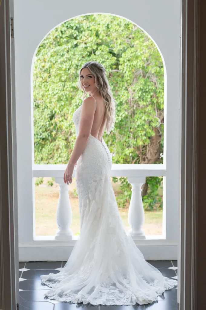 Arthur-Andrea-Jamaica-Halfmoon-Wedding-189.jpg - OMOS Photography Bride in a white lace wedding gown stands in an arched window, smiling at the camera with a green garden backdrop behind her.