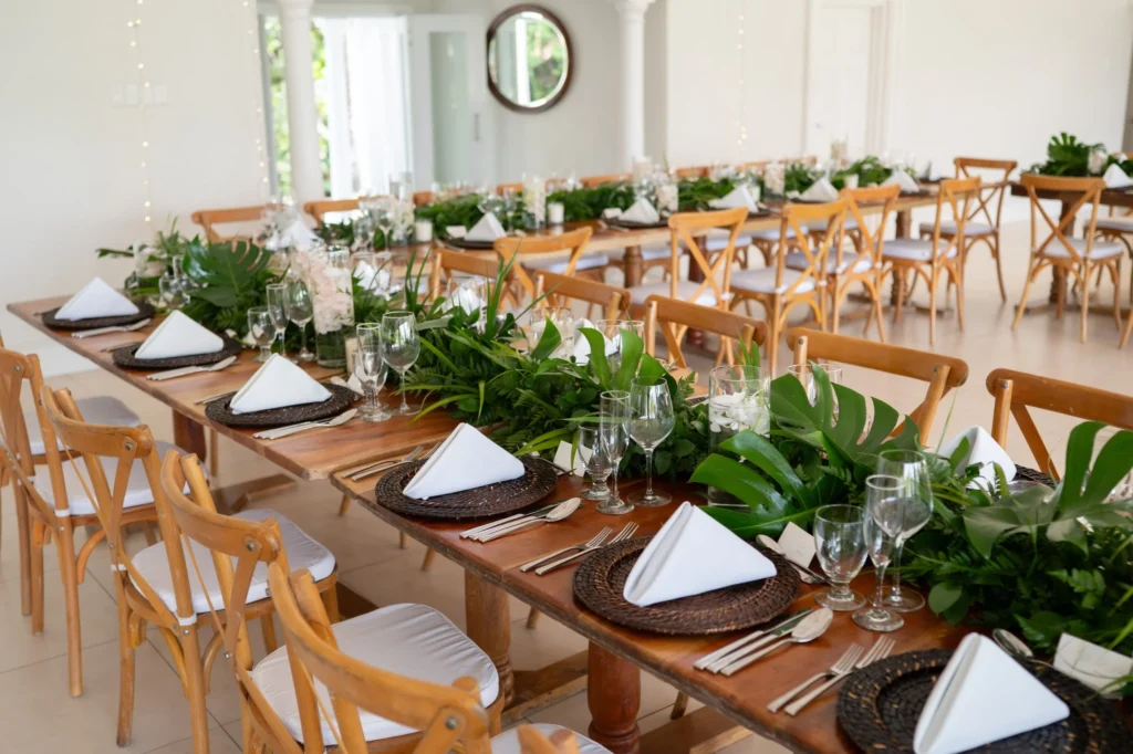 Arthur-Andrea-Jamaica-Halfmoon-Wedding-210.jpg - OMOS Photography Long wooden dining tables arranged for a formal event with white napkins, glassware, and lush green centerpieces in a bright room.