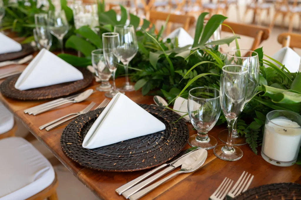 Arthur-Andrea-Jamaica-Halfmoon-Wedding-211.jpg - OMOS Photography Table setting with woven placemats, white napkins folded beside silverware and clear glasses, greenery centerpiece A close-up of an elegant dining table with napkins, forks and wine glasses amid leafy greenery Chic table setup featuring white napkins, multiple glasses, and a lush green garland down the center Wooden table laid for guests: napkins, silverware, glassware, and a verdant plant centerpiece Formal dining scene focusing on place settings and fresh greenery accents