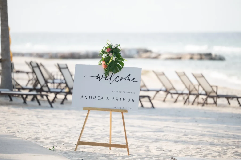 Arthur-Andrea-Jamaica-Halfmoon-Wedding-214.jpg - OMOS Photography Beach wedding welcome sign on an easel with a floral bouquet, set among empty chairs by the shore.