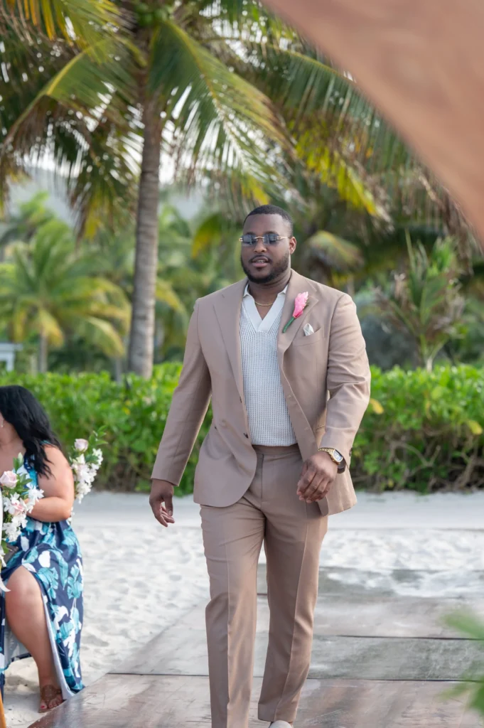 Arthur-Andrea-Jamaica-Halfmoon-Wedding-228.jpg - OMOS Photography Man in a beige suit with a pink flower boutonniere and sunglasses standing outdoors on a sandy path with palm trees in the background; tropical setting.