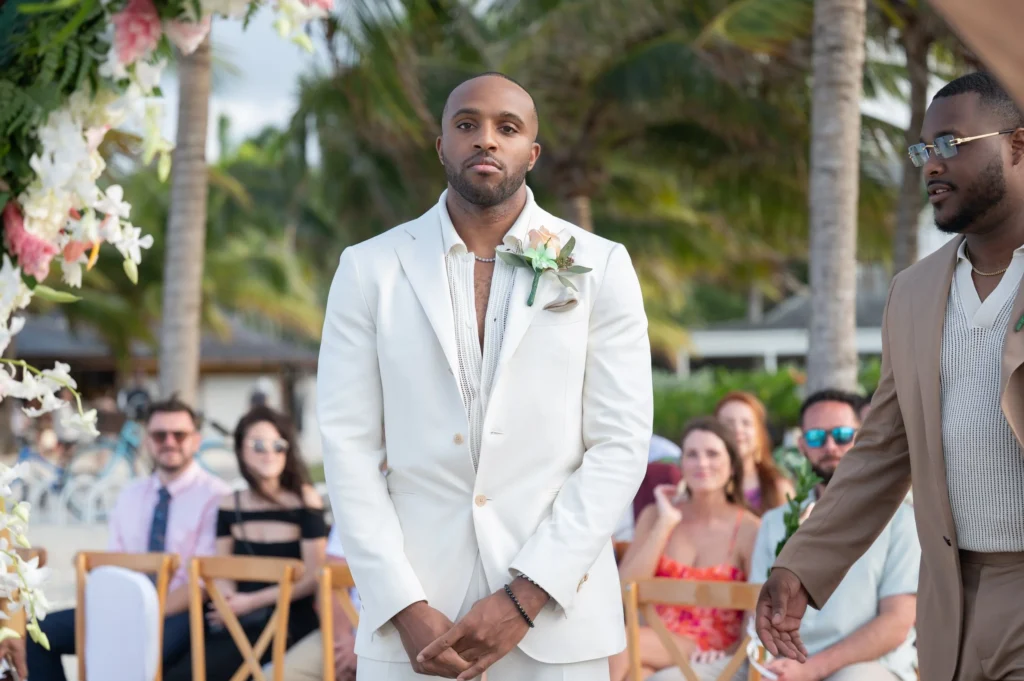 Arthur-Andrea-Jamaica-Halfmoon-Wedding-229.jpg - OMOS Photography Groom in a white suit with a boutonniere standing at an outdoor wedding ceremony, guests seated behind under palm trees.
