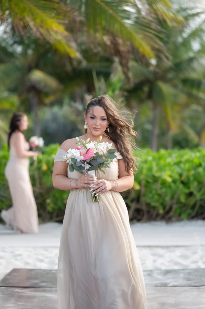 Arthur-Andrea-Jamaica-Halfmoon-Wedding-235.jpg - OMOS Photography Bride in a beige off-shoulder gown holding a pink-and-white bouquet on a sunny beach, with a bridesmaid in the background.