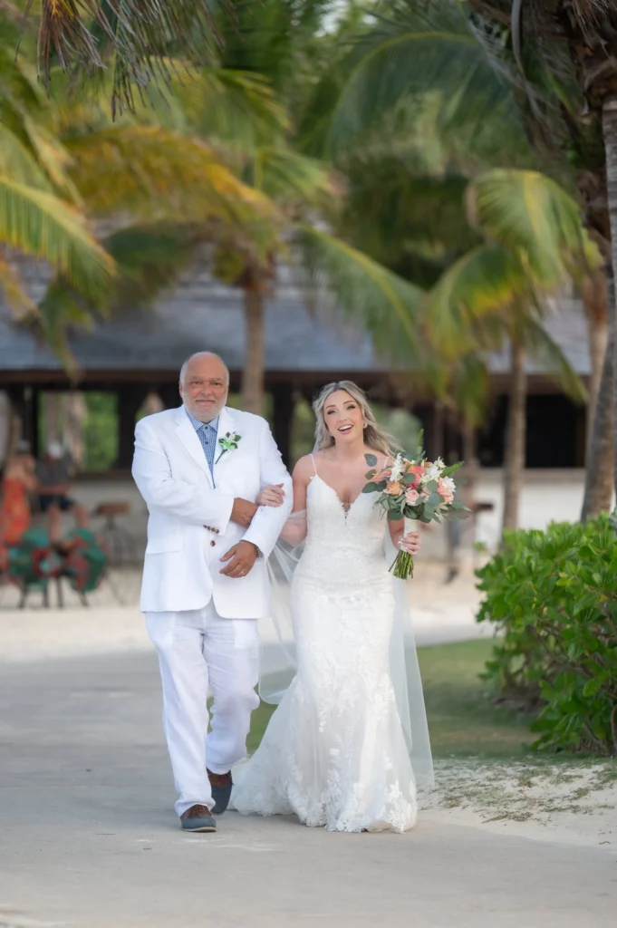 Arthur-Andrea-Jamaica-Halfmoon-Wedding-252.jpg - OMOS Photography An older man in a white suit walks arm-in-arm with a smiling bride in a lace white wedding gown, holding a bouquet, along a palm-fringed path outdoors.