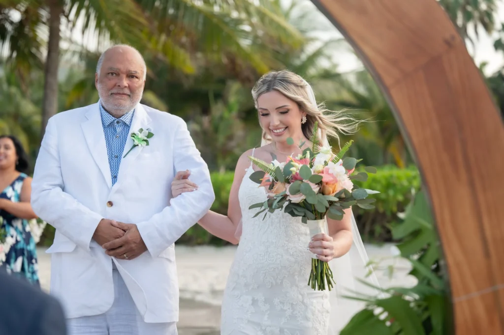 Arthur-Andrea-Jamaica-Halfmoon-Wedding-256.jpg - OMOS Photography Bride in a lace wedding dress holds a bouquet while an older man in a white suit escorts her outdoors beside a wooden arch and tropical plants.
