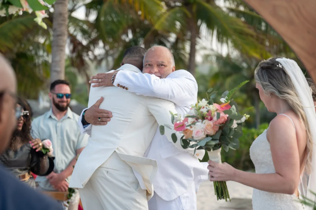 Arthur-Andrea-Jamaica-Halfmoon-Wedding-258.jpg - OMOS Photography Two men in white suits share a warm hug at an outdoor tropical wedding, with the bride in a white dress and a colorful bouquet nearby.