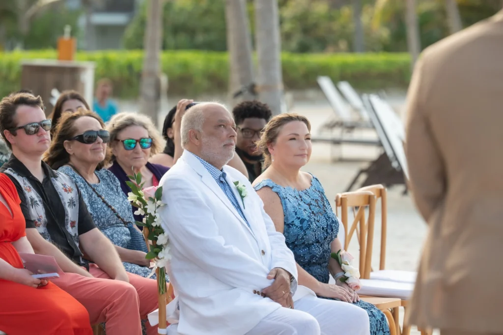 Arthur-Andrea-Jamaica-Halfmoon-Wedding-268.jpg - OMOS Photography Group of wedding guests seated along a beach ceremony, some holding flowers, with palm trees in the background.