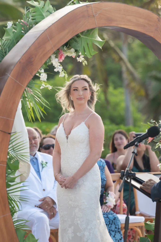 Arthur-Andrea-Jamaica-Halfmoon-Wedding-272.jpg - OMOS Photography Bride in a white lace gown stands under a curved wooden arch decorated with tropical leaves and pink/white flowers at an outdoor ceremony with guests in the background.