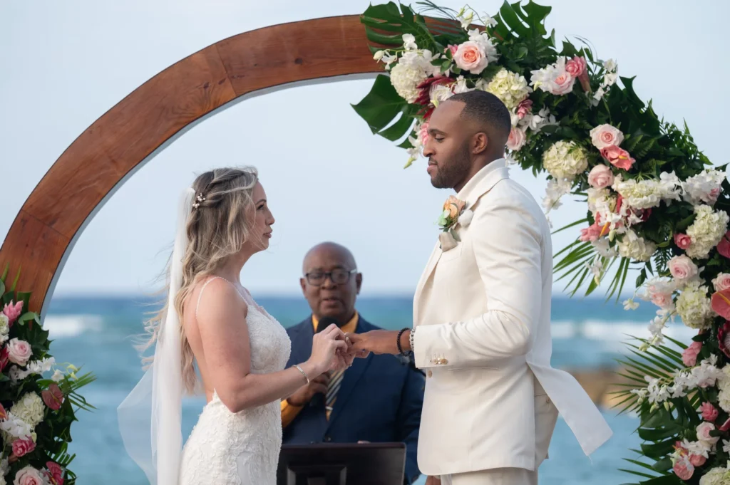 Arthur-Andrea-Jamaica-Halfmoon-Wedding-287.jpg - OMOS Photography Bride and groom exchange rings under a flower-covered arch on the beach, officiant in the background.