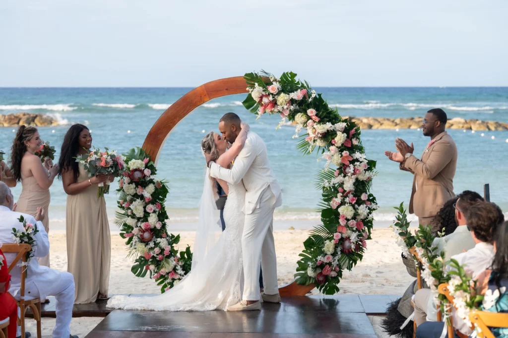 Arthur-Andrea-Jamaica-Halfmoon-Wedding-297.jpg - OMOS Photography Bride and groom kiss under a circular floral arch on a sunny beach, with bridesmaids and guests nearby by the shore.