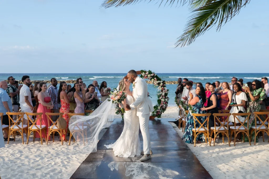 Arthur-Andrea-Jamaica-Halfmoon-Wedding-304.jpg - OMOS Photography Bride and groom kiss under a floral arch on a sunny beach with seated guests along a wooden aisle by the sea.