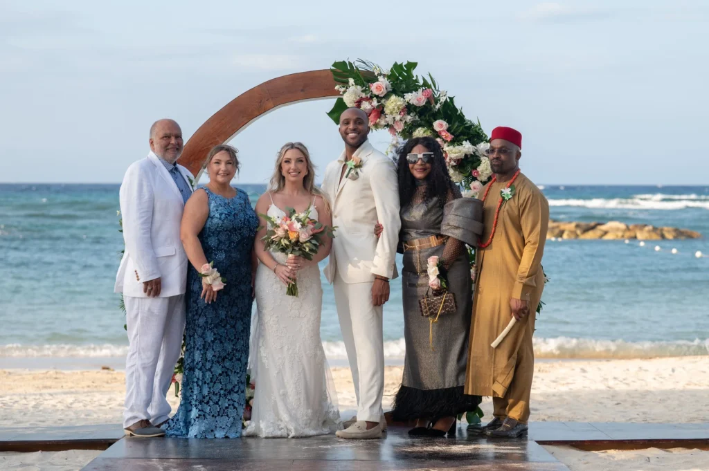 Arthur-Andrea-Jamaica-Halfmoon-Wedding-325.jpg - OMOS Photography Six people in wedding attire pose on a sandy beach with a large flower arch and ocean behind them.