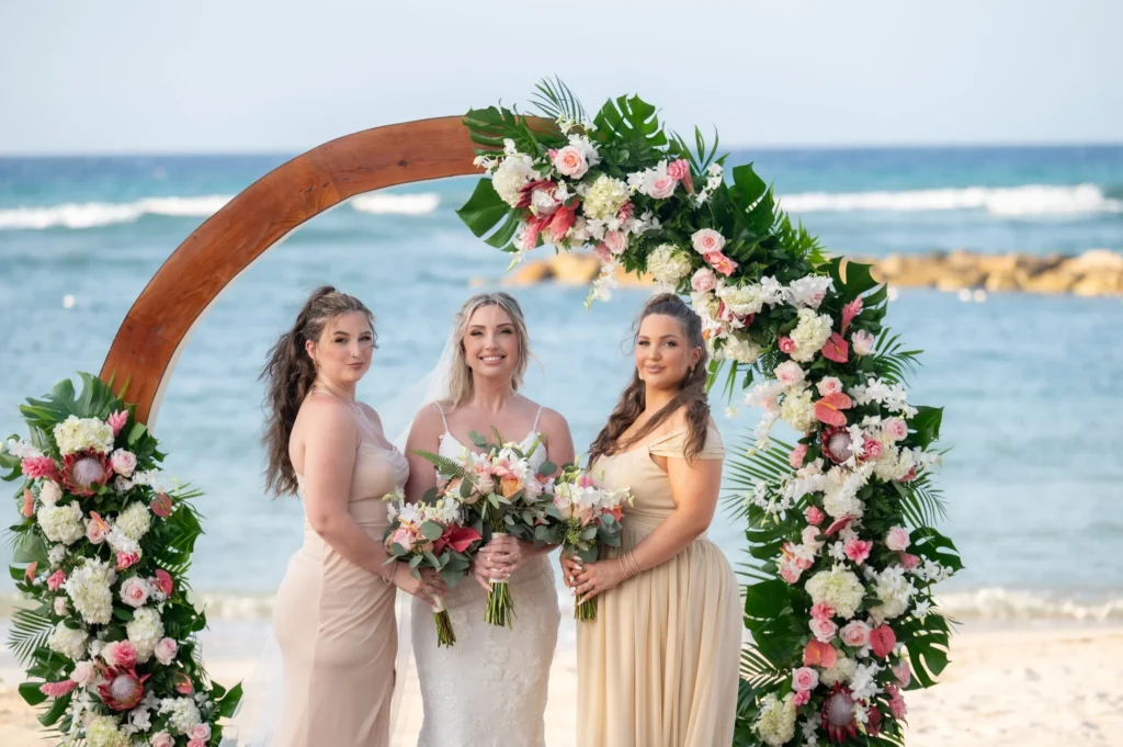 Arthur-Andrea-Jamaica-Halfmoon-Wedding-336.jpg - OMOS Photography Bride in a white wedding gown flanked by two bridesmaids in beige dresses, standing under a large floral arch on a sunny beach with the ocean in the background.