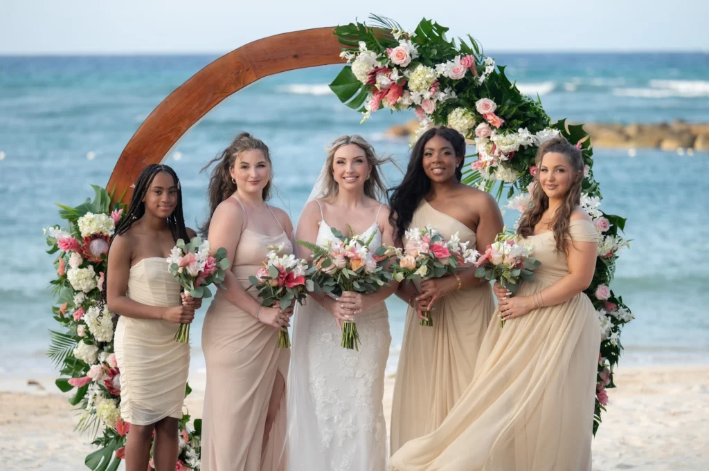 Arthur-Andrea-Jamaica-Halfmoon-Wedding-339.jpg - OMOS Photography Bride and five bridesmaids in beige dresses pose with a floral arch on a sunny beach by the sea, holding bouquets.