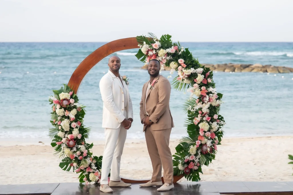 Arthur-Andrea-Jamaica-Halfmoon-Wedding-352.jpg - OMOS Photography Two men in light-colored suits stand hand in hand under a floral arch on a beach, with the sea behind them.