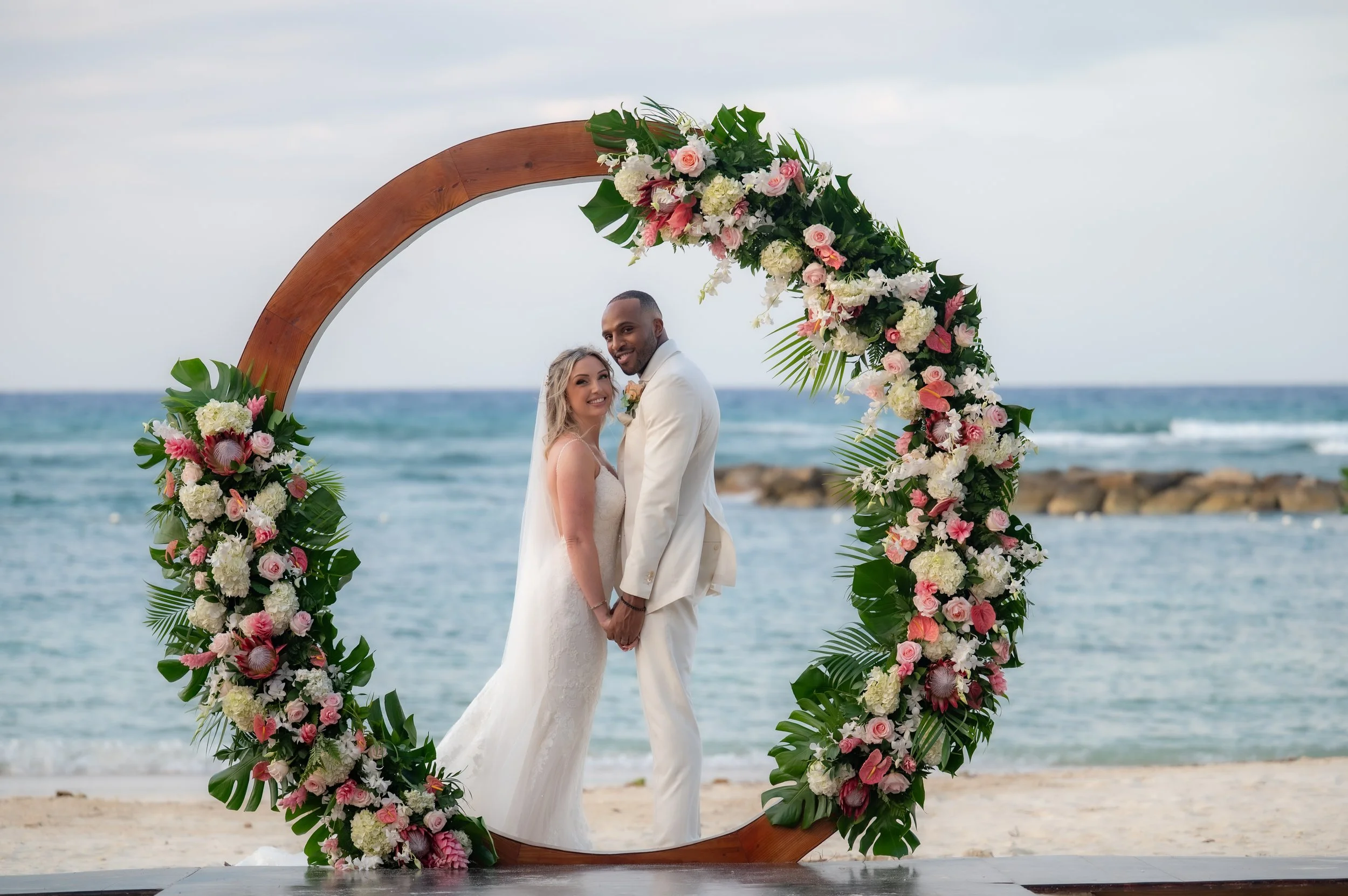Bride and groom standing hand in hand under a circular flower arch on the beach, with the ocean in the background.