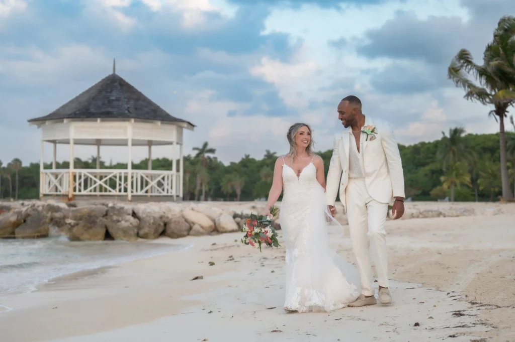 Arthur-Andrea-Jamaica-Halfmoon-Wedding-376.jpg - OMOS Photography A couple in wedding attire walks hand in hand along a sandy beach near a thatched gazebo with palm trees in the background.