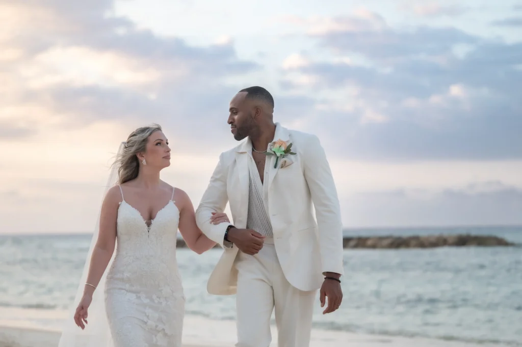 Arthur-Andrea-Jamaica-Halfmoon-Wedding-389.jpg - OMOS Photography Bride in a lace wedding gown and groom in a light suit walk hand in hand along the beach at sunset with a pastel sky behind them.