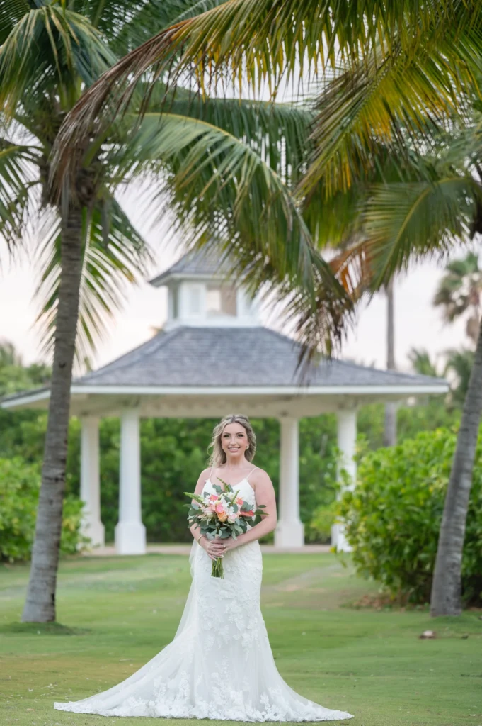 Arthur-Andrea-Jamaica-Halfmoon-Wedding-399.jpg - OMOS Photography Bride in a white lace wedding gown standing on a lawn with a bouquet, palm trees and a gazebo in the background.