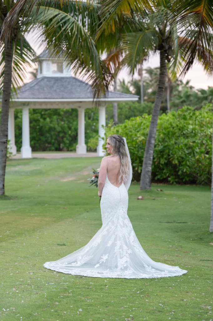 Arthur-Andrea-Jamaica-Halfmoon-Wedding-414.jpg - OMOS Photography Bride in a white lace wedding gown with a long veil, seen from the back, standing on a green lawn with palm trees and a pavilion in the background.