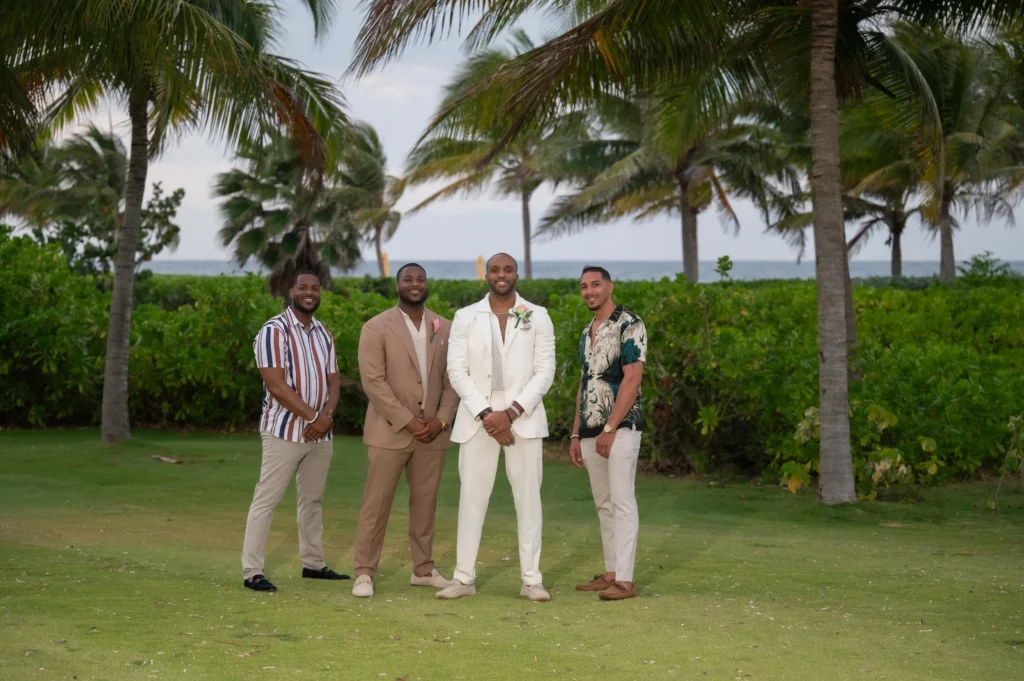 Arthur-Andrea-Jamaica-Halfmoon-Wedding-426.jpg - OMOS Photography Four men stand together on a grassy lawn with palm trees and the ocean in the background, posing for a group photo.