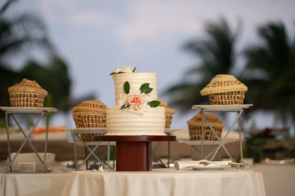 Arthur-Andrea-Jamaica-Halfmoon-Wedding-430.jpg - OMOS Photography Center-tiered white wedding cake with pink rose and green leaves on a wooden stand, outdoors with wicker baskets in the background.