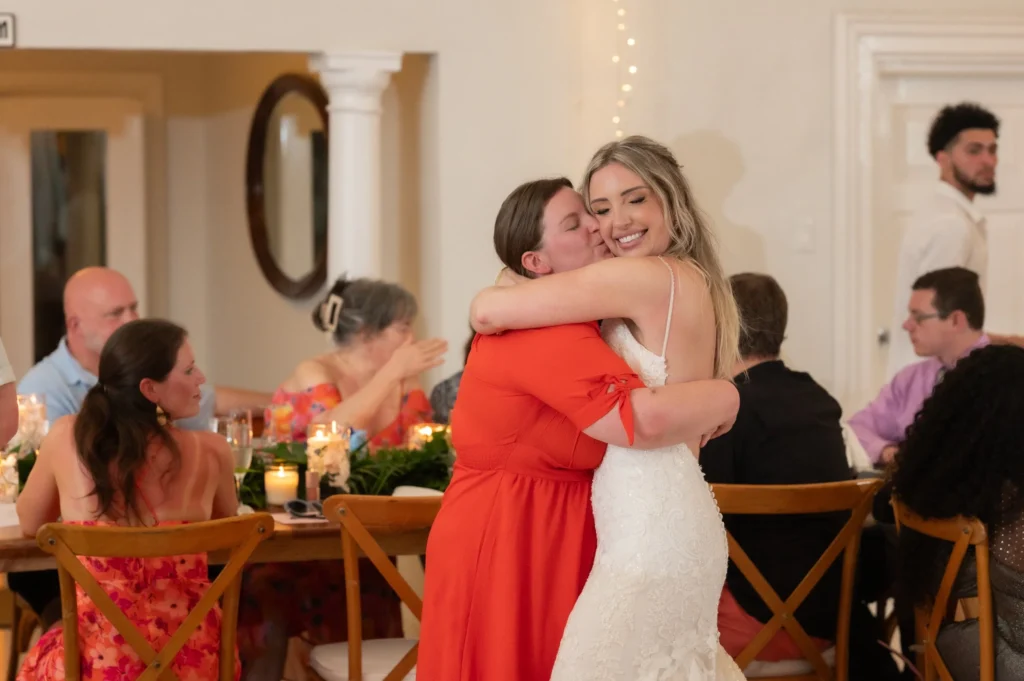 Arthur-Andrea-Jamaica-Halfmoon-Wedding-504.jpg - OMOS Photography Bride in a white wedding dress hugs a woman in a bright red dress at a wedding reception, smiling with candlelit tables in the background.