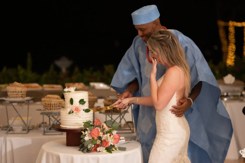 Arthur-Andrea-Jamaica-Halfmoon-Wedding-514.jpg - OMOS Photography Bride in a white lace dress and a man in blue traditional attire cut a wedding cake at night outdoors, with flowers in the foreground.