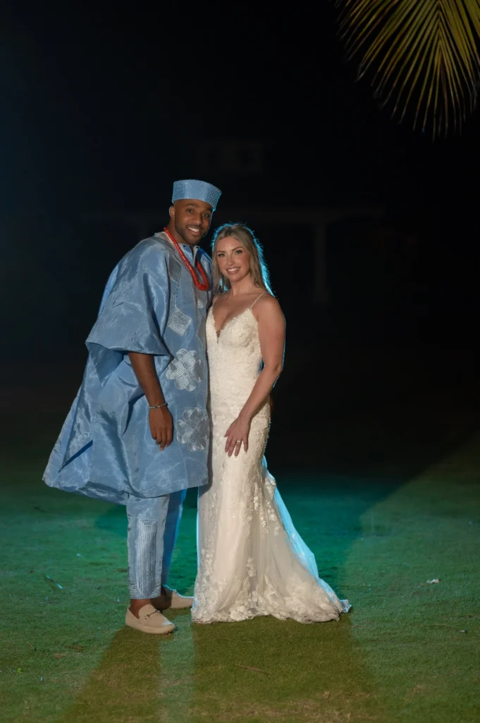 Arthur-Andrea-Jamaica-Halfmoon-Wedding-529.jpg - OMOS Photography Nighttime outdoor photo of a couple: man in blue traditional attire with matching hat and a bride in a white lace wedding dress, standing on grass.