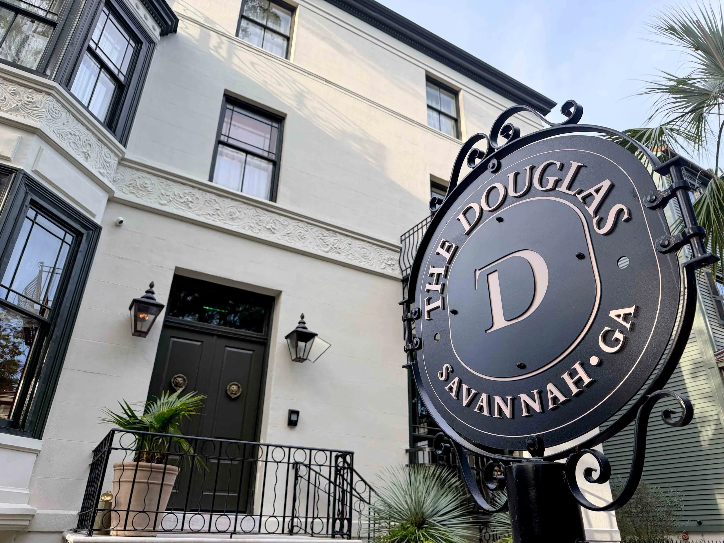 White, multi-story building with black-framed windows and decorative floral molding along the facade, seen beside a large circular metal sign reading 'DOUGLAS' in the foreground.