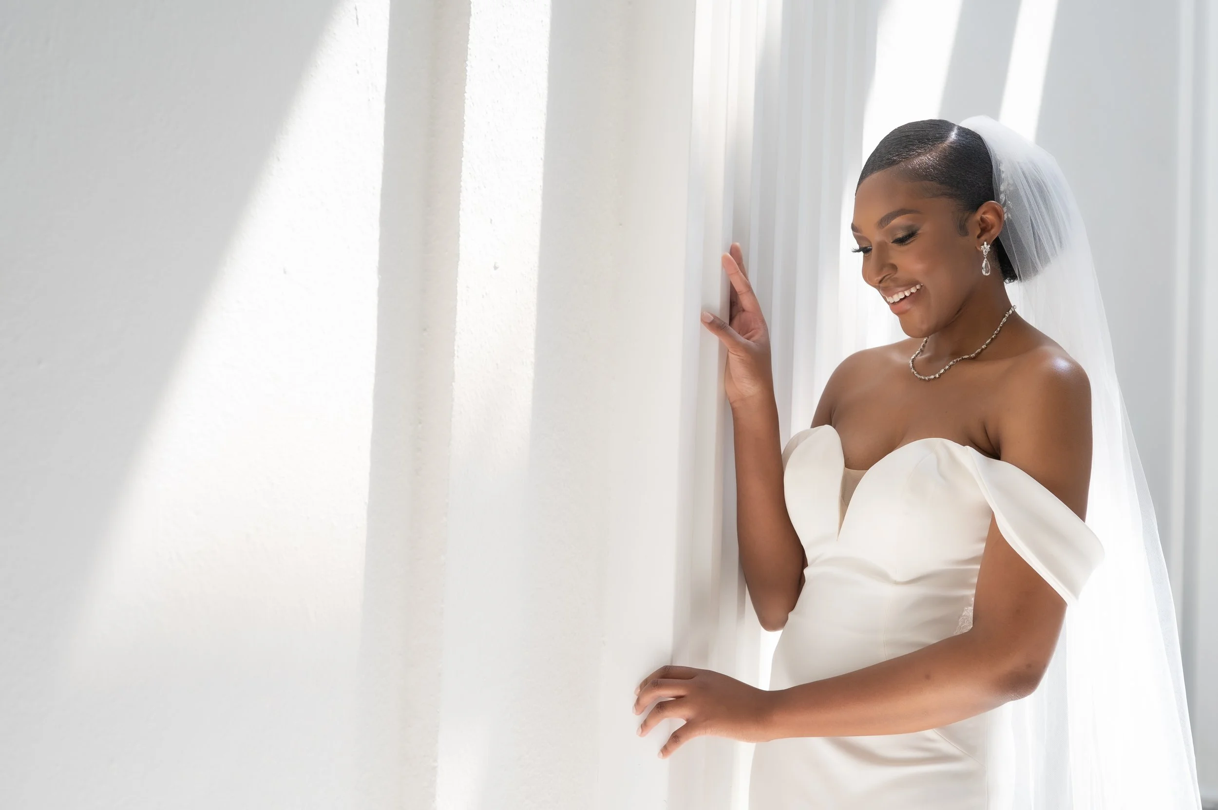 Bride in a white off-shoulder wedding gown with veil and jewelry smiles beside white draped curtains.