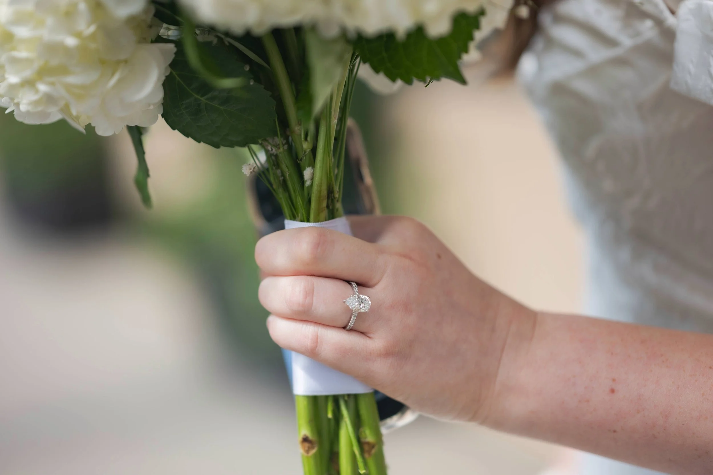 Close-up of a hand holding a white-flower bouquet with green stems, tied with a white ribbon, wearing a diamond engagement ring and another band.