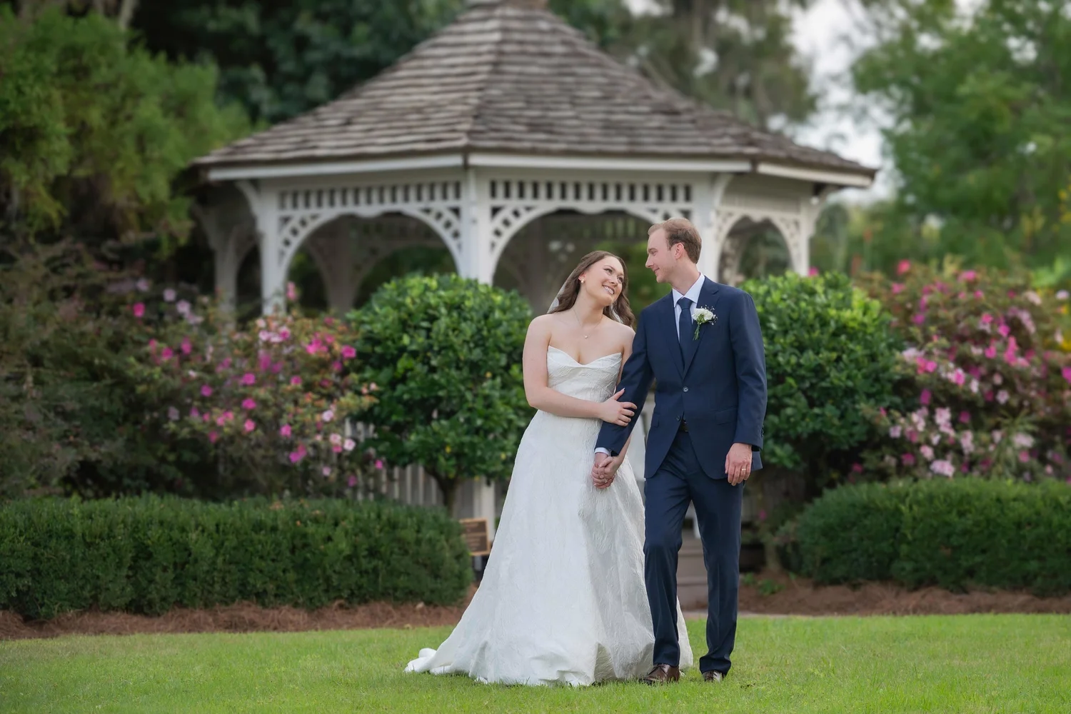 Bride in a white wedding gown and groom in a navy suit walk hand in hand in a garden with a gazebo behind them.
