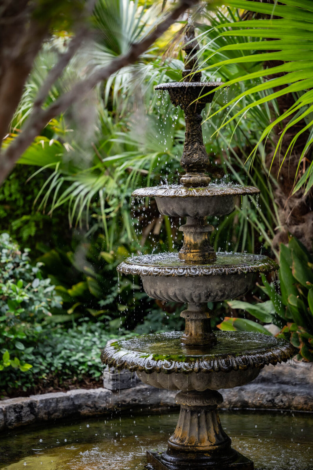 Ornate three-tier stone fountain in a lush tropical garden with water dripping from each tier