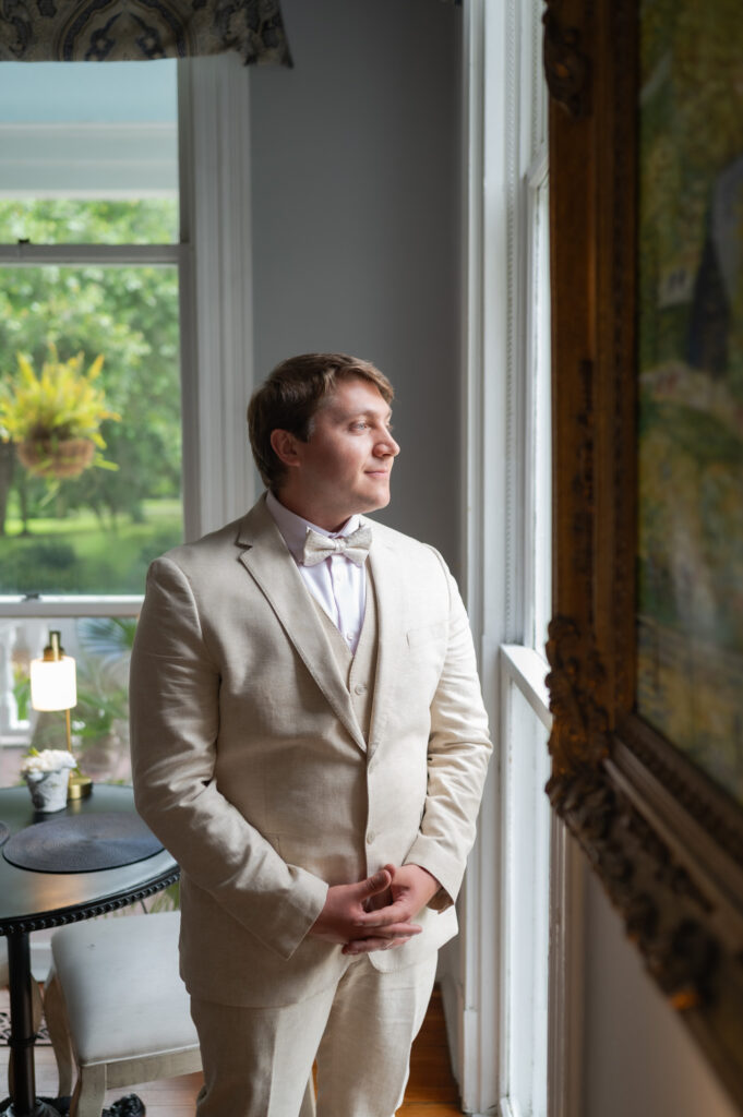 Man in a beige suit with a bow tie standing by a window, looking contemplative outside the window.