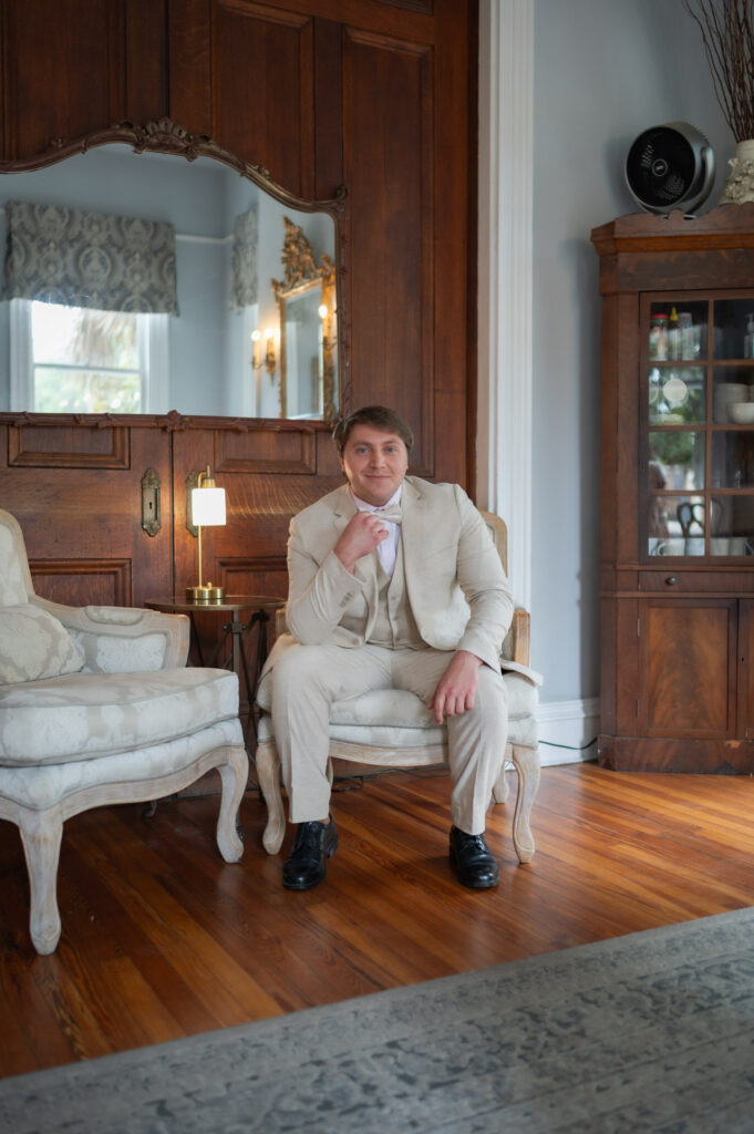 Man in a beige suit sits on a patterned chair in a wood-paneled, elegant living room.