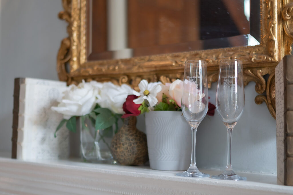 Decorative mantel display with a gold ornate frame, white and red flowers, and two tall champagne flutes in the foreground.