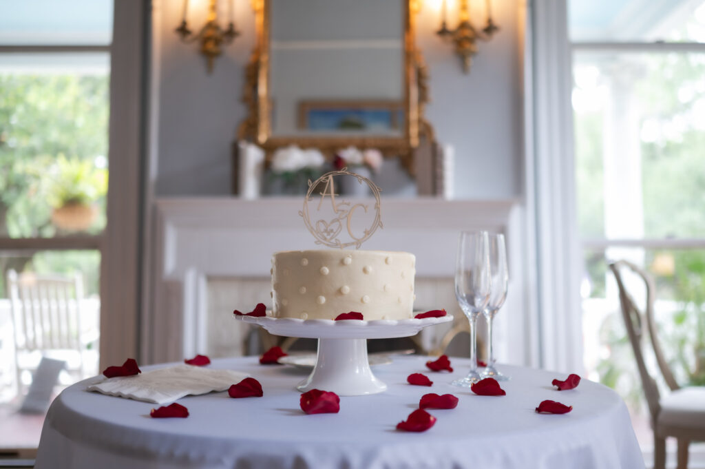 White frosted cake with pearl-like decorations on a pedestal, red rose petals scattered on a white tablecloth in the foreground.