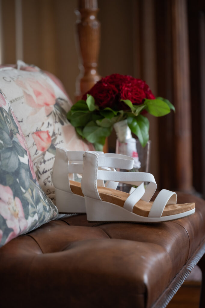 White strappy platform sandals resting on a brown leather couch, with a floral pillow and a red bouquet visible in the background.