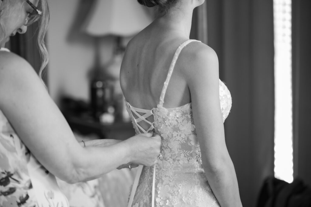 Bride in a lace wedding dress being laced up from behind by a helper, shown from the back side in black and white.