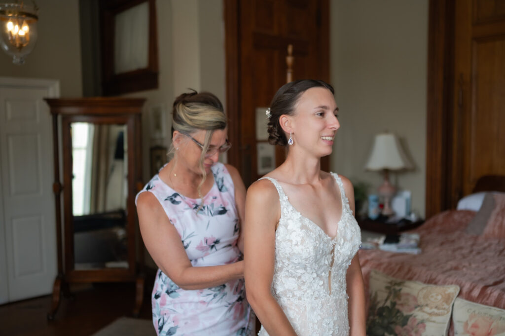 Bride in a lace wedding gown stands as a woman in a floral dress helps adjust the back of her dress in a cozy room.
