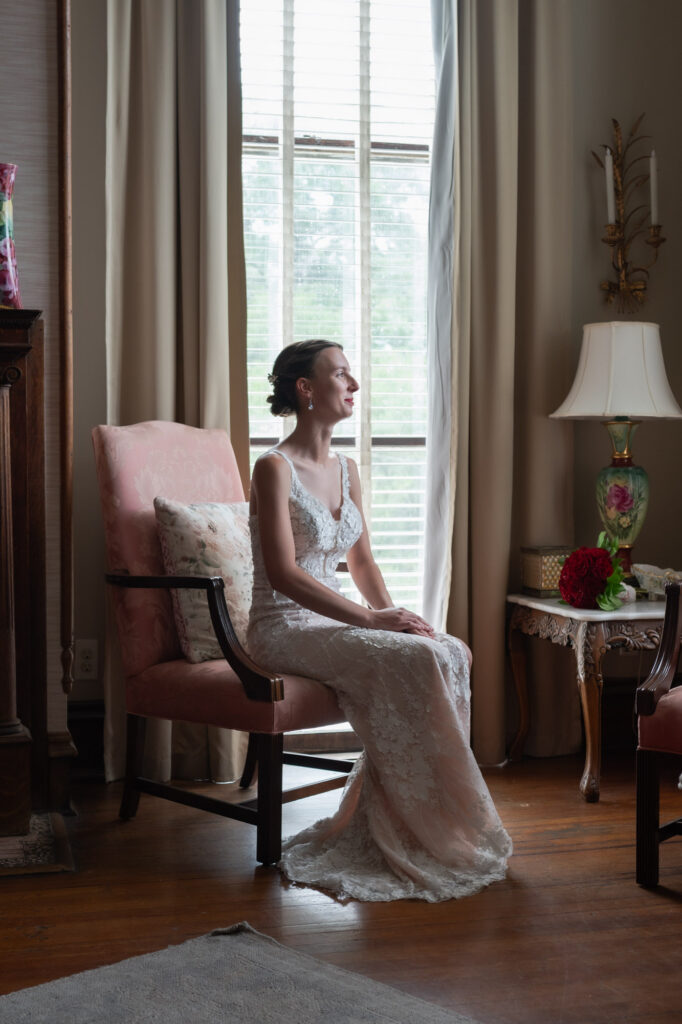 Bride in a white lace wedding dress sits gracefully in a pink upholstered chair by a sunlit window, hands folded in her lap, with a bouquet of red roses on a table nearby.