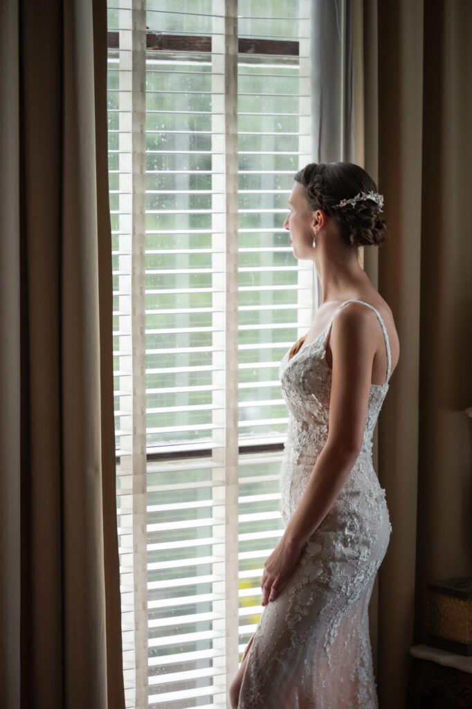 Bride in a lace wedding gown stands by a window, gazing outside with a soft, natural light highlighting her updo and floral hairpiece.